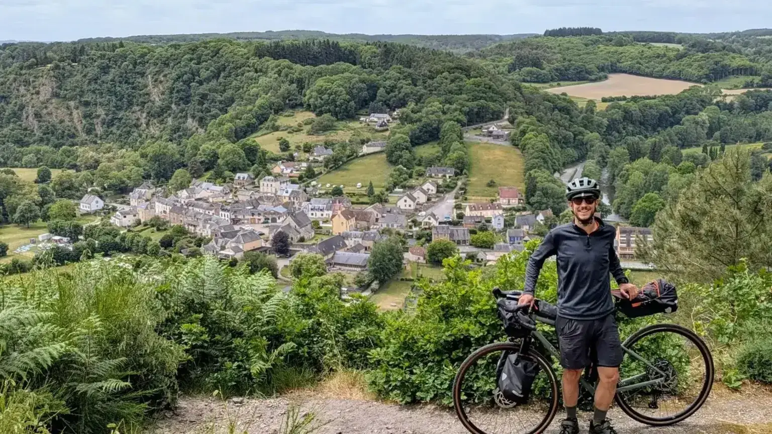 Cyclotouriste et son vélo chargé au-dessus d'un village niché dans une vallée boisée